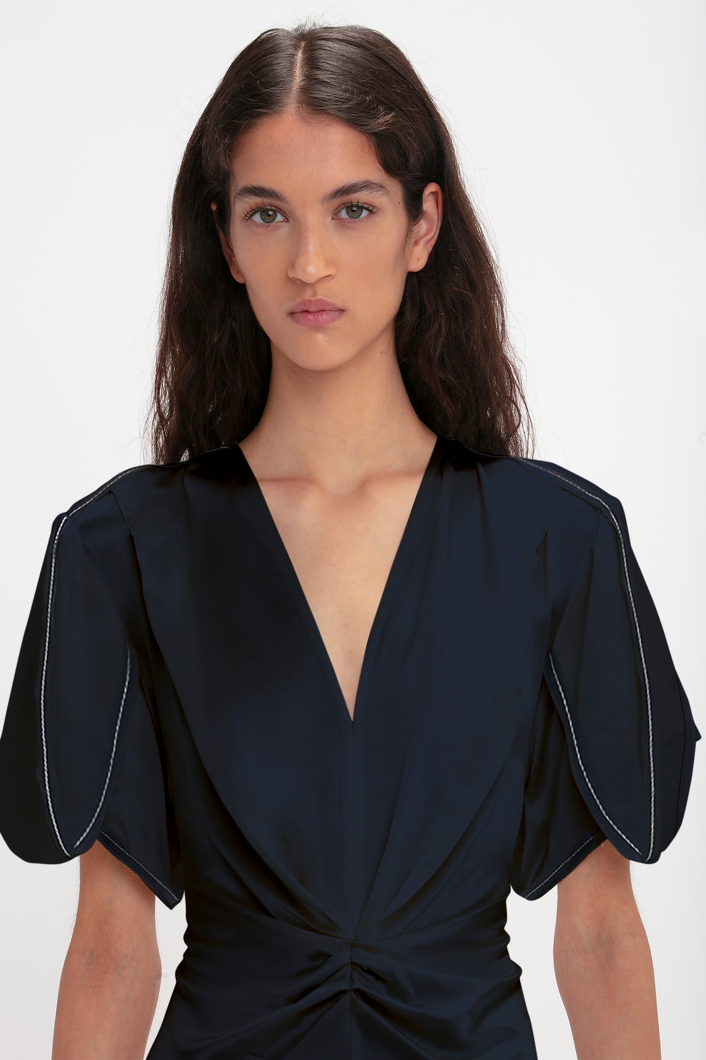 A woman with shoulder-length brown hair, wearing a dark Victoria Beckham Exclusive Gathered V-Neck Midi Dress In Navy with puffed sleeves, looking directly at the camera.