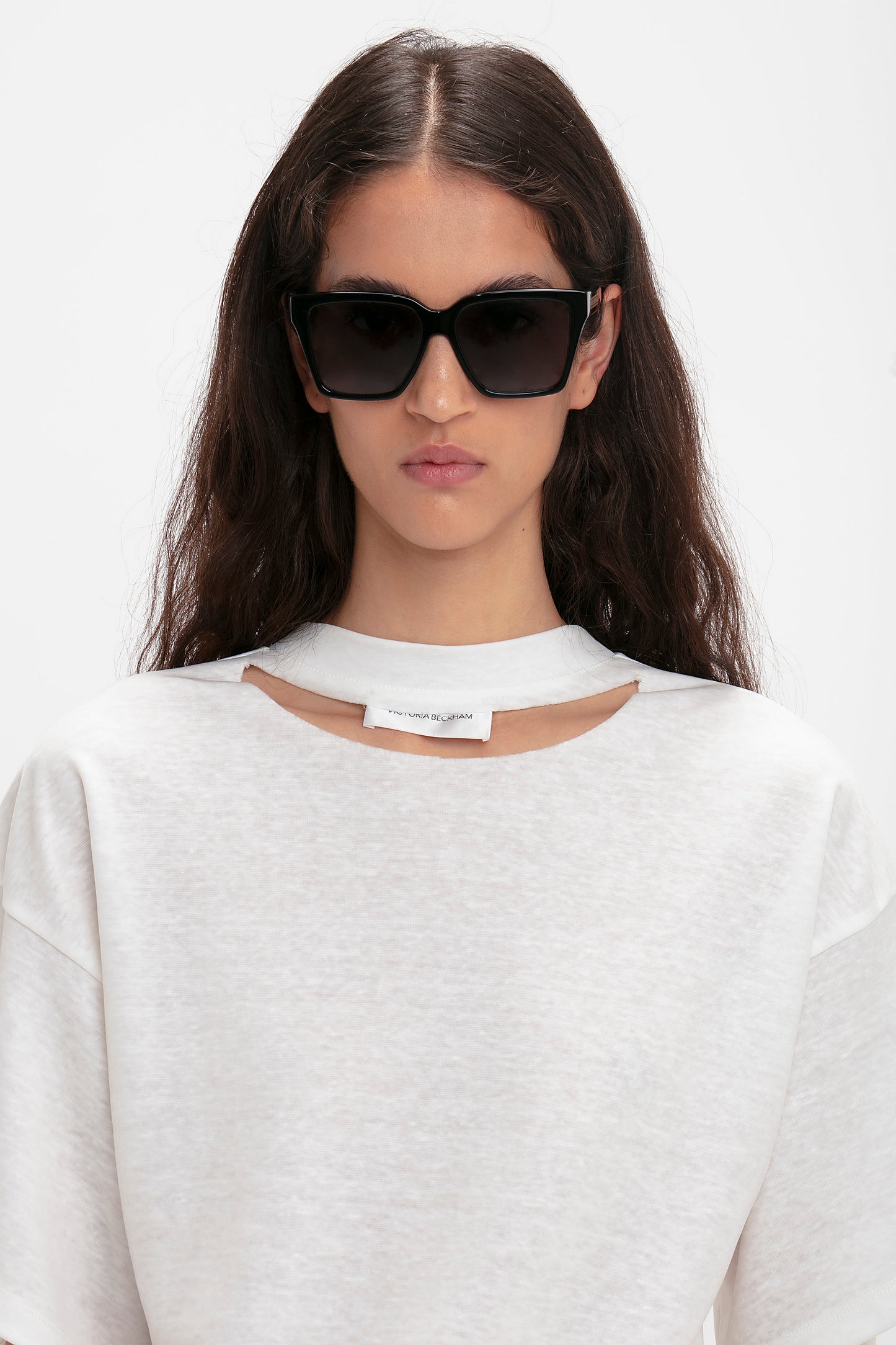 Wearing Victoria Beckham eyewear, a person with long dark hair sports large Soft Square Frame Sunglasses In Black and a white top with a cut-out detail and a tag on the collar.
