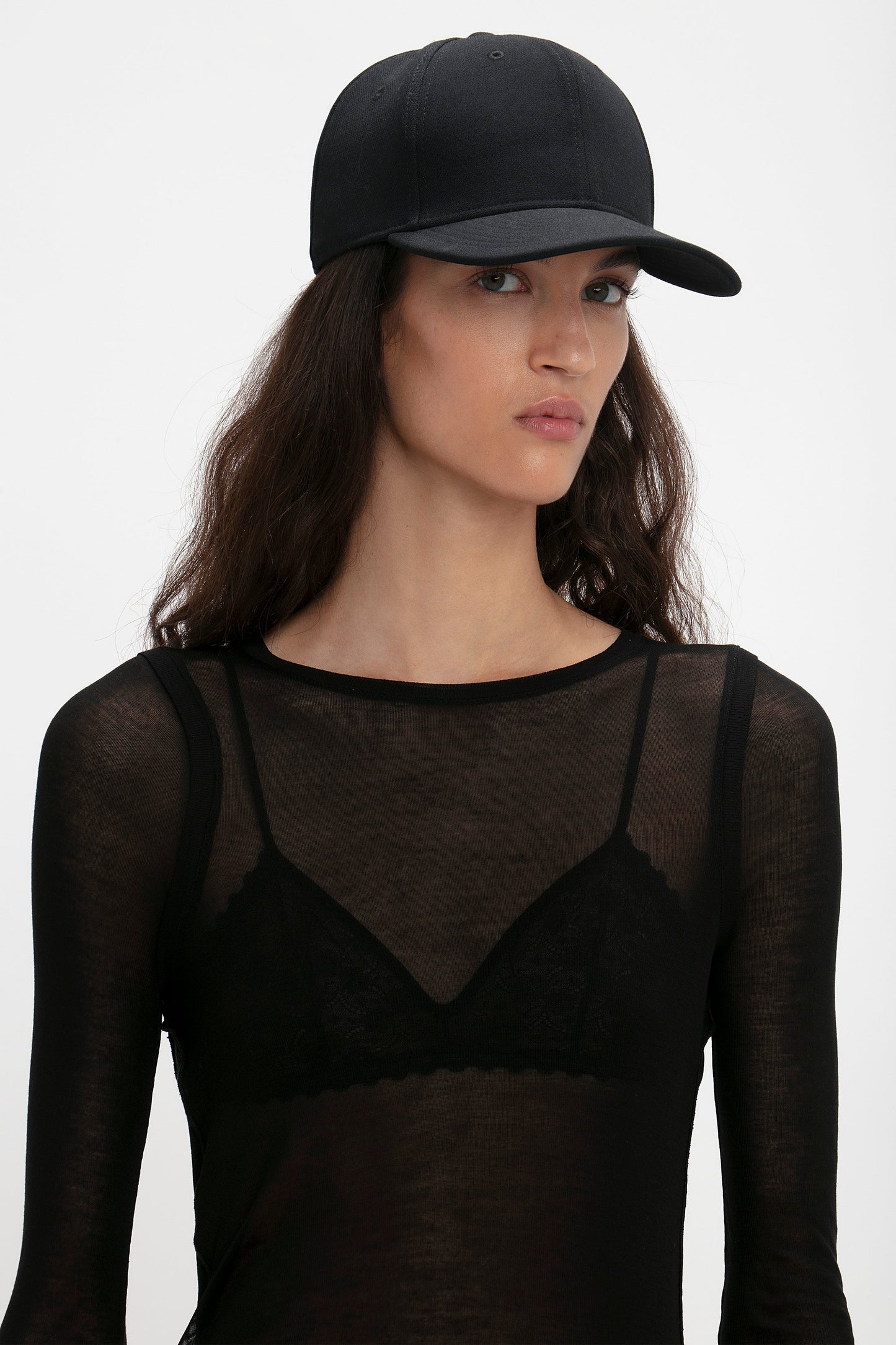 A woman wearing a Victoria Beckham Exclusive Logo Cap In Black and a sheer black top with lace detailing, looking toward the camera.