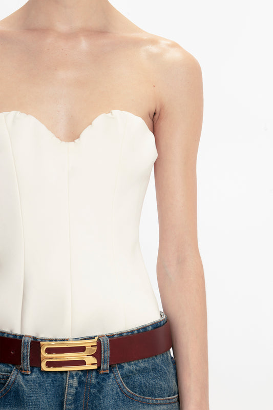 Close-up of a woman wearing a Victoria Beckham antique white corset top and blue jeans with a brown belt featuring a golden buckle, set against a white background.