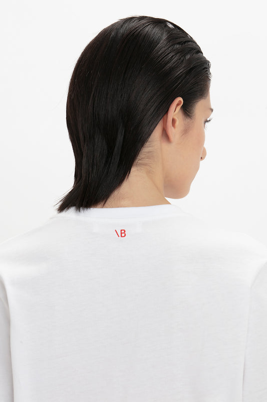 Rear view of a person with shoulder-length dark hair, wearing a Victoria Beckham 'David's Wife' Slogan T-Shirt In White with a small red logo on the collar.
