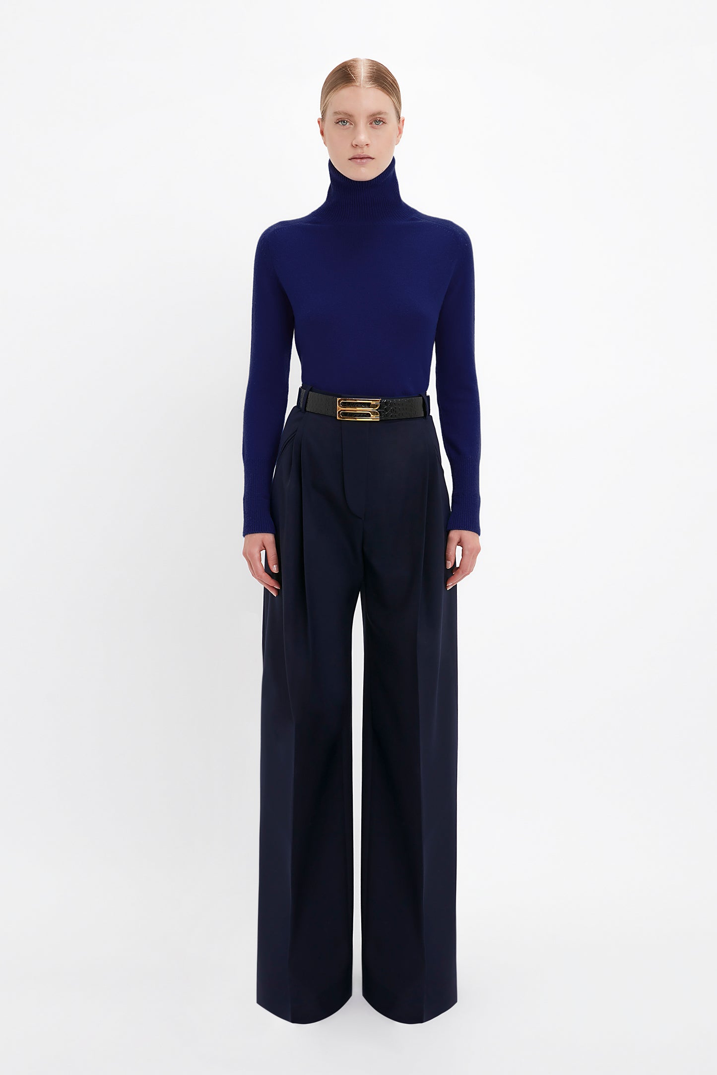 Person wearing a Victoria Beckham Polo Neck Jumper In Navy with a chic saddle shoulder design and high-waisted, wide-leg trousers with a black belt, standing against a plain white background.