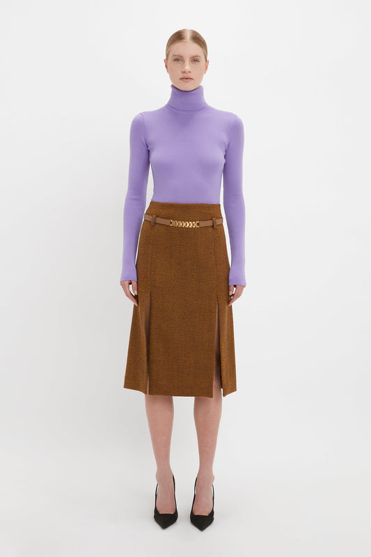 A woman in a purple turtleneck and brown skirt stands against a plain white background, wearing black Pointy Toe Pumps by Victoria Beckham.