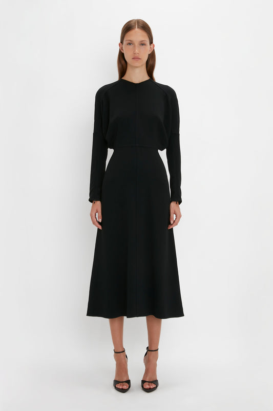 A woman in a black Victoria Beckham Dolman Midi Dress with long sleeves and squared toe sandals stands against a white background.