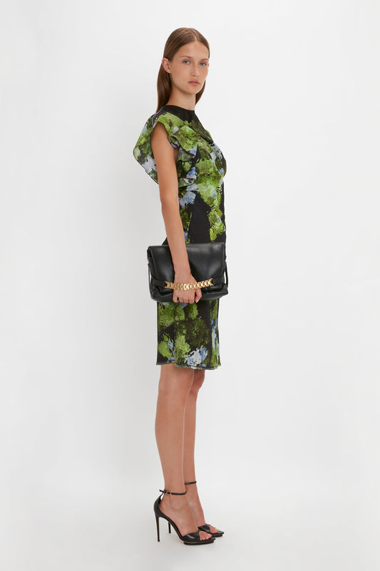 A woman models a green and blue floral dress with black heels and carries a Victoria Beckham Puffy Chain Pouch With Strap In Black Leather, posing in a plain white studio setting.