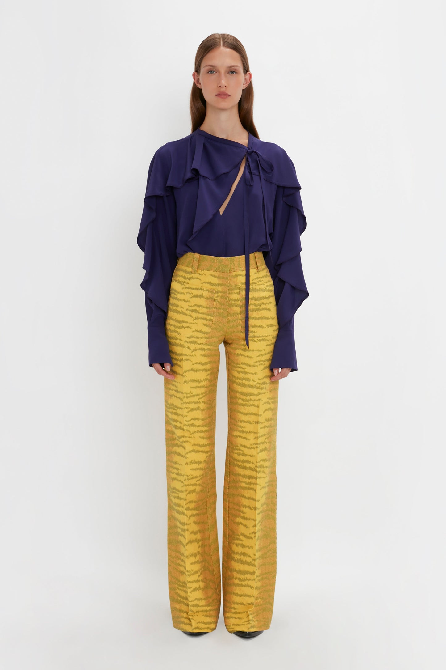 A person stands against a white background, wearing the Tie Detail Ruffle Blouse In Ultraviolet by Victoria Beckham with long sleeves and yellow high-waisted, wide-leg pants with an abstract pattern.