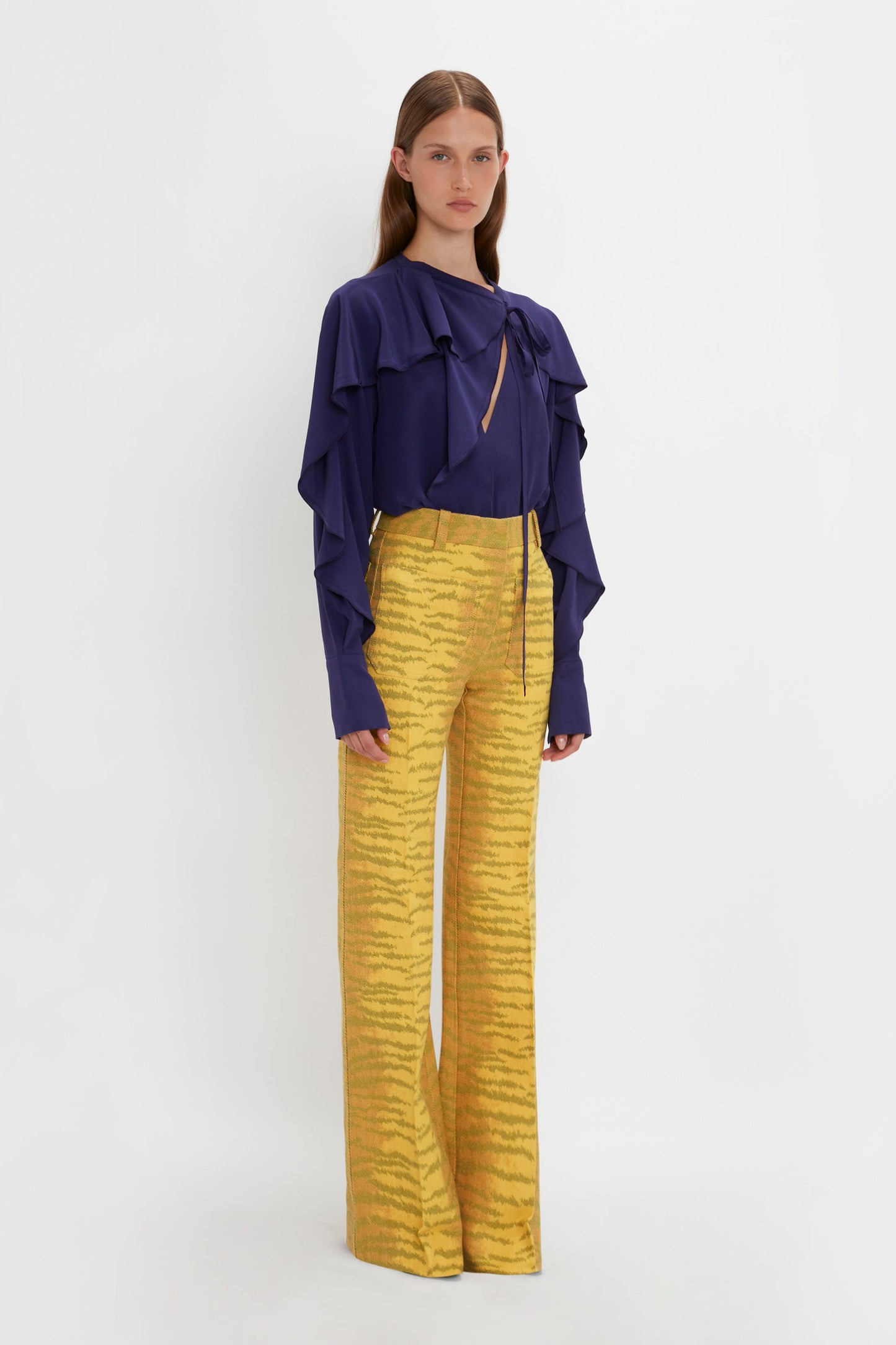Person in yellow tiger-striped wide-leg pants and a dark blue Tie Detail Ruffle Blouse In Ultraviolet by Victoria Beckham stands against a white background.