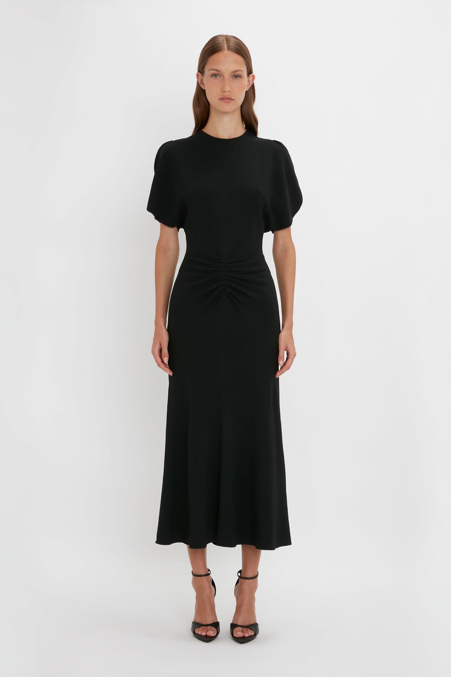 A young woman wearing a black Gathered Waist Midi Dress by Victoria Beckham, with a twisted front detail and short sleeves, standing against a plain white background.