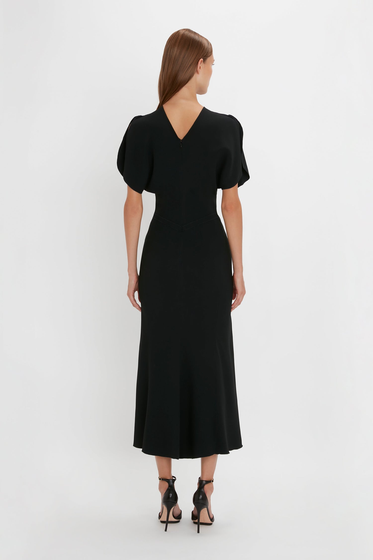 Woman standing with her back to the camera, wearing a Victoria Beckham Gathered Waist Midi Dress in Black with short sleeves and v-back, paired with black pointy toe stilettos on a white backdrop.
