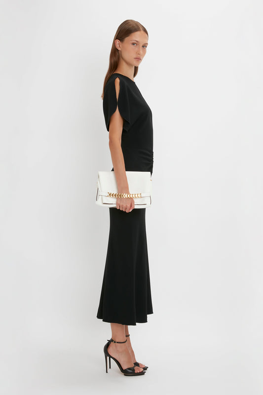 Woman in a black Victoria Beckham fit-and-flare silhouette dress and pointy toe stiletto heels holding a white handbag, standing sideways against a white background.