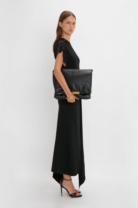 A woman in a Victoria Beckham Short Sleeve Tie Detail Dress In Black and heels holds a large black clutch with a golden chain, posing side-on against a white background.