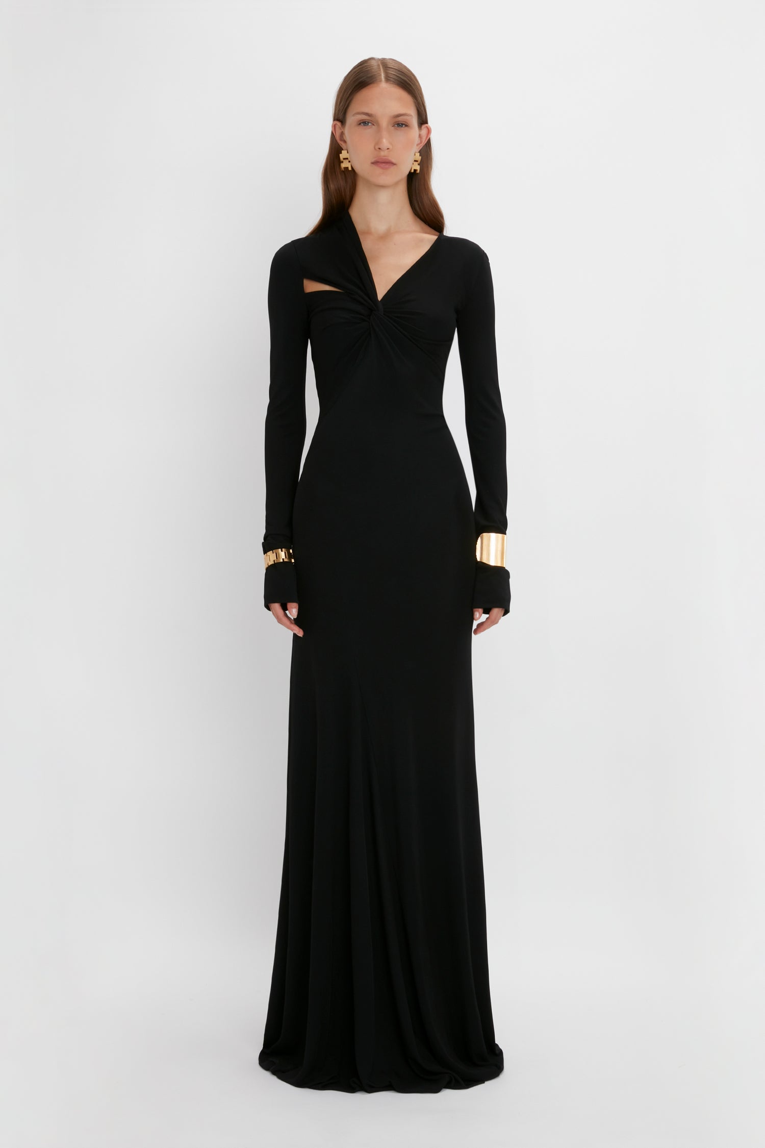 A woman stands against a plain white background, wearing a long black dress with long sleeves and a twisted front detail, accessorized with Exclusive Perfume Cuff bracelets in gold-plated brass and earrings by Victoria Beckham.