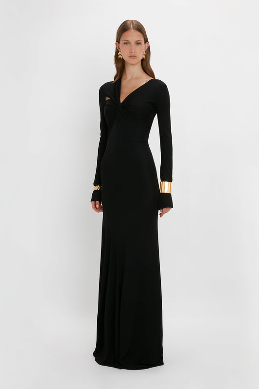 Woman in a Victoria Beckham Tie Detail Floor-Length Dress in Black with gold cuffs, standing against a white background.