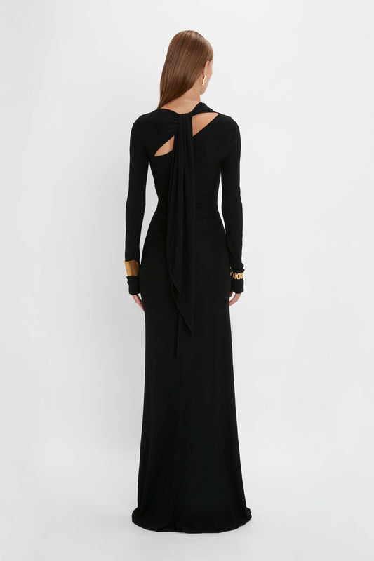 A woman in a Victoria Beckham Tie Detail Floor-Length Dress in Black with a draped back and gold cuffs, standing against a white background.