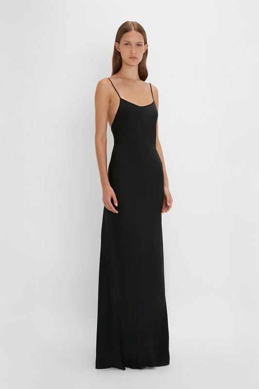 A person stands facing the camera, wearing a Floor-Length Cami Dress In Black by Victoria Beckham made of crepe back satin with thin straps. The background is plain and white.