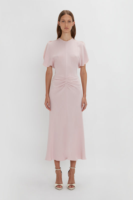 A woman in a Victoria Beckham pale pink gathered waist midi dress with tulip sleeves and a knot detail at the waist, standing against a white background.