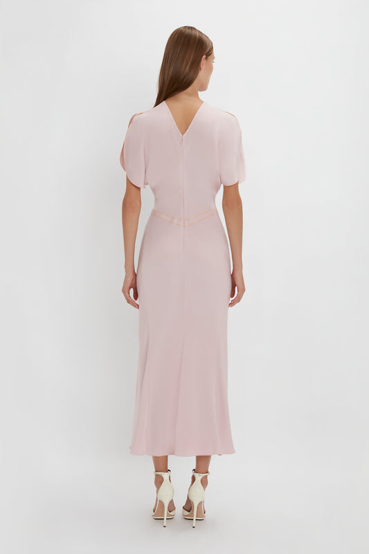 A woman stands facing away, wearing a Victoria Beckham Gathered Waist Midi Dress In Blush with tulip sleeves and white heels, against a plain white background.