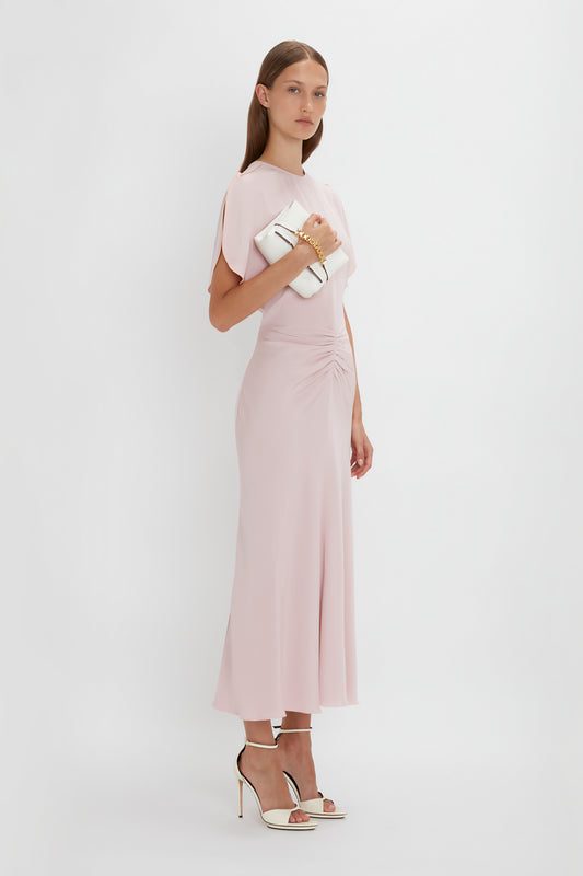 A woman in a Victoria Beckham Gathered Waist Midi Dress In Blush with tulip sleeves, standing with one hand on her hip, holding a white clutch, and wearing strappy heels against a white background.