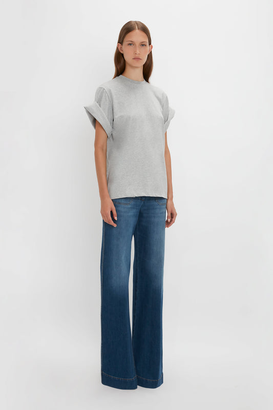 A woman standing against a white background wearing a grey Asymmetric Relaxed Fit T-Shirt In Grey Marl and blue flared jeans by Victoria Beckham.