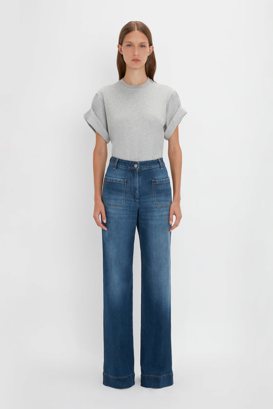 A woman stands against a white background, wearing a gray t-shirt and blue high-waisted Victoria Beckham Alina jeans in Dark Vintage Wash.