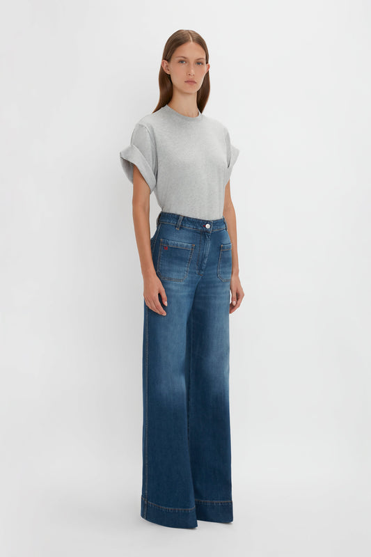 A woman wearing a gray t-shirt and blue high-waisted, wide-leg Victoria Beckham Alina Jeans in Dark Vintage Wash standing against a white background.
