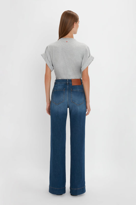 A woman stands with her back to the camera, wearing a Victoria Beckham asymmetric relaxed fit t-shirt in grey marl made of organic cotton and blue high-waisted jeans with a brown belt.