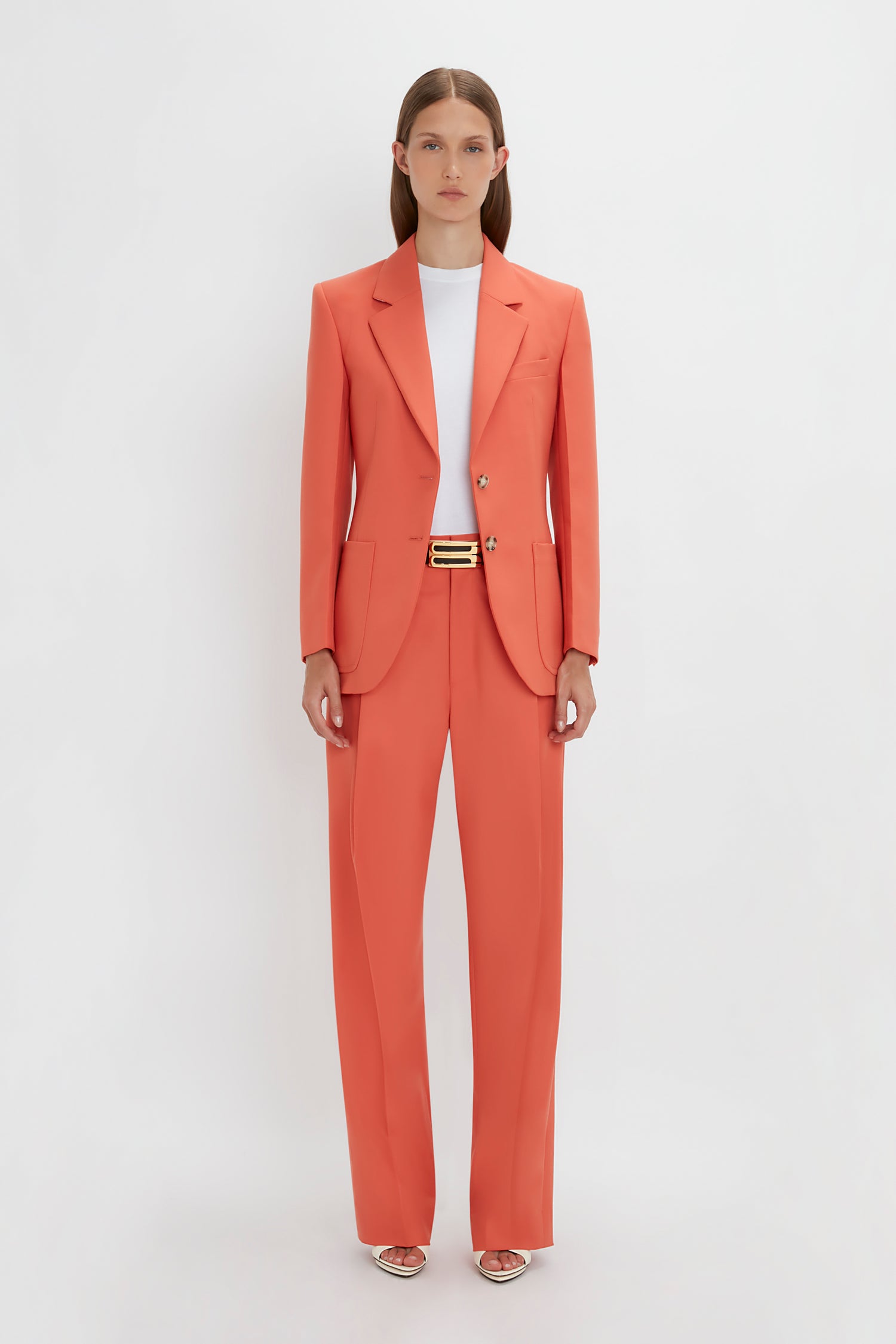 A woman in a Victoria Beckham Patch Pocket Jacket In Papaya with straight-leg trousers and a matching blazer, accessorized with a black belt, stands against a white background.