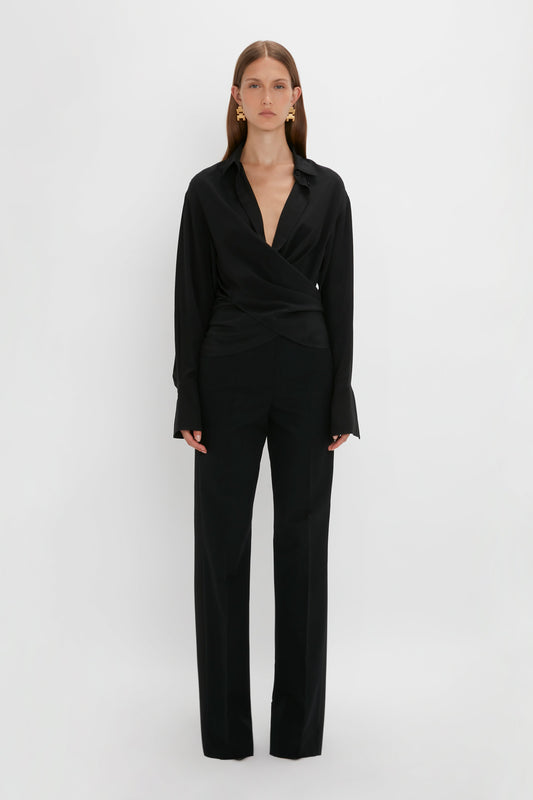 A woman stands against a white background, wearing a tailored black Victoria Beckham wrap blazer and matching oversized fit trousers.