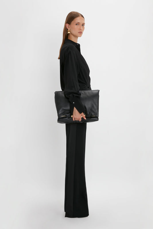 A woman stands in profile, wearing a Victoria Beckham black silk wrap front blouse and black trousers with a large black tote bag, against a white background.