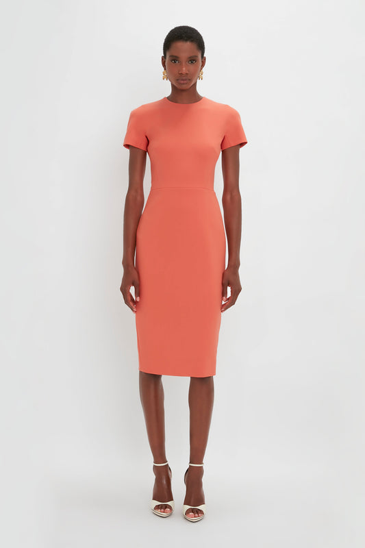 A woman standing in a studio, wearing a Victoria Beckham orange midi-length dress with short sleeves and open-toe heels, looking at the camera.