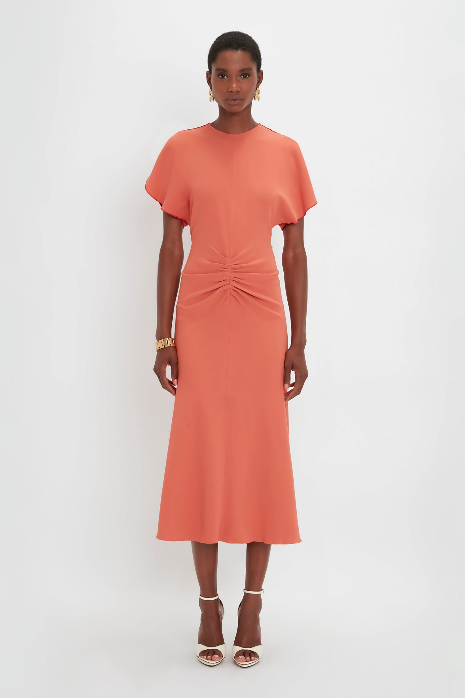 A woman standing against a white background, wearing an orange midi dress with short sleeves and a twisted detail at the waist, paired with Pointy Toe Stiletto heels. She is wearing the Gathered Waist Midi Dress In Papaya by Victoria Beckham.