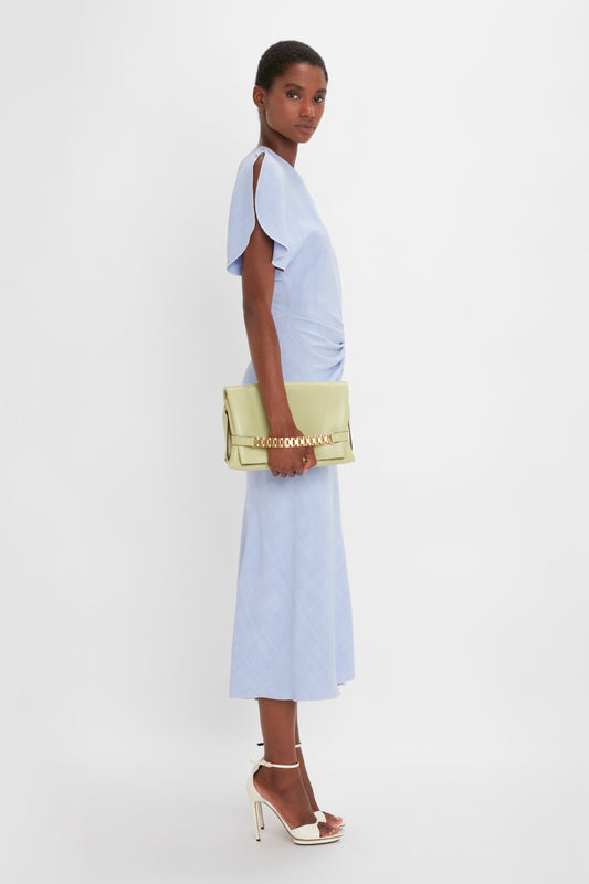 A woman in a light blue dress and white heels holds a Victoria Beckham Chain Pouch With Strap In Avocado Leather handbag, posed against a white background.