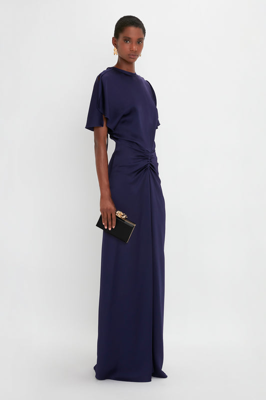 A woman in a refined navy evening gown with a twisted detail at the waist, holding a small Victoria Beckham Frame Flower Minaudiere in Black clutch with a gold flower clasp.