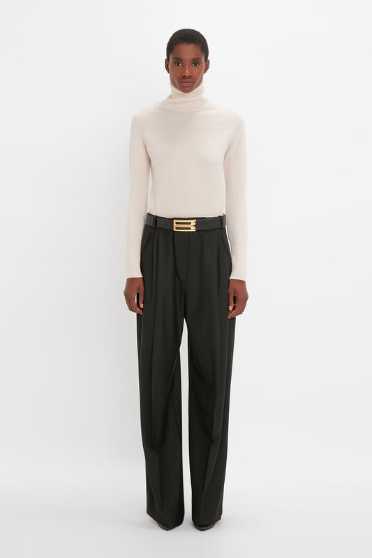 A woman stands against a white background wearing a light beige lambswool sweater and black trousers with a gold buckle belt featuring the Victoria Beckham monogram.