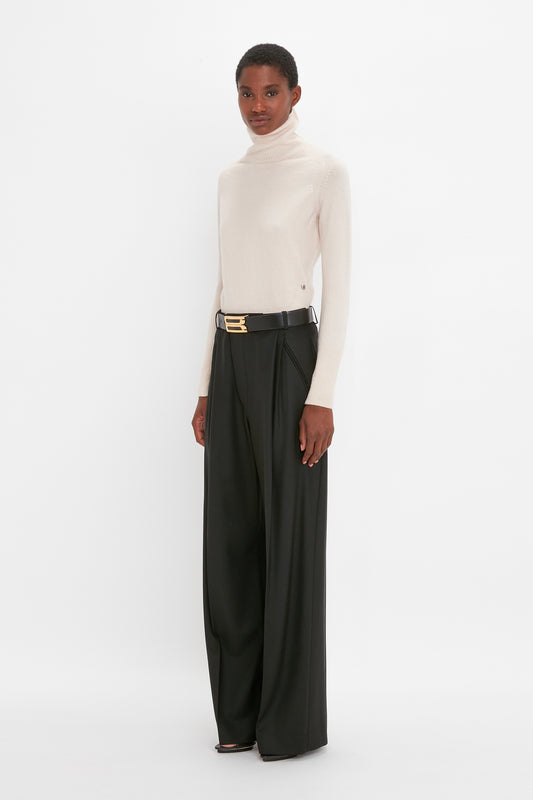 A woman in a Victoria Beckham ivory Polo Neck Jumper and black trousers with a gold belt stands against a plain background.