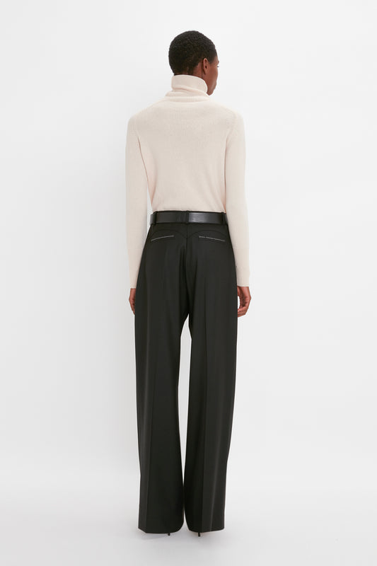 A woman stands facing away, wearing a Victoria Beckham high-neck lambswool sweater and black wide-leg trousers against a plain white background.