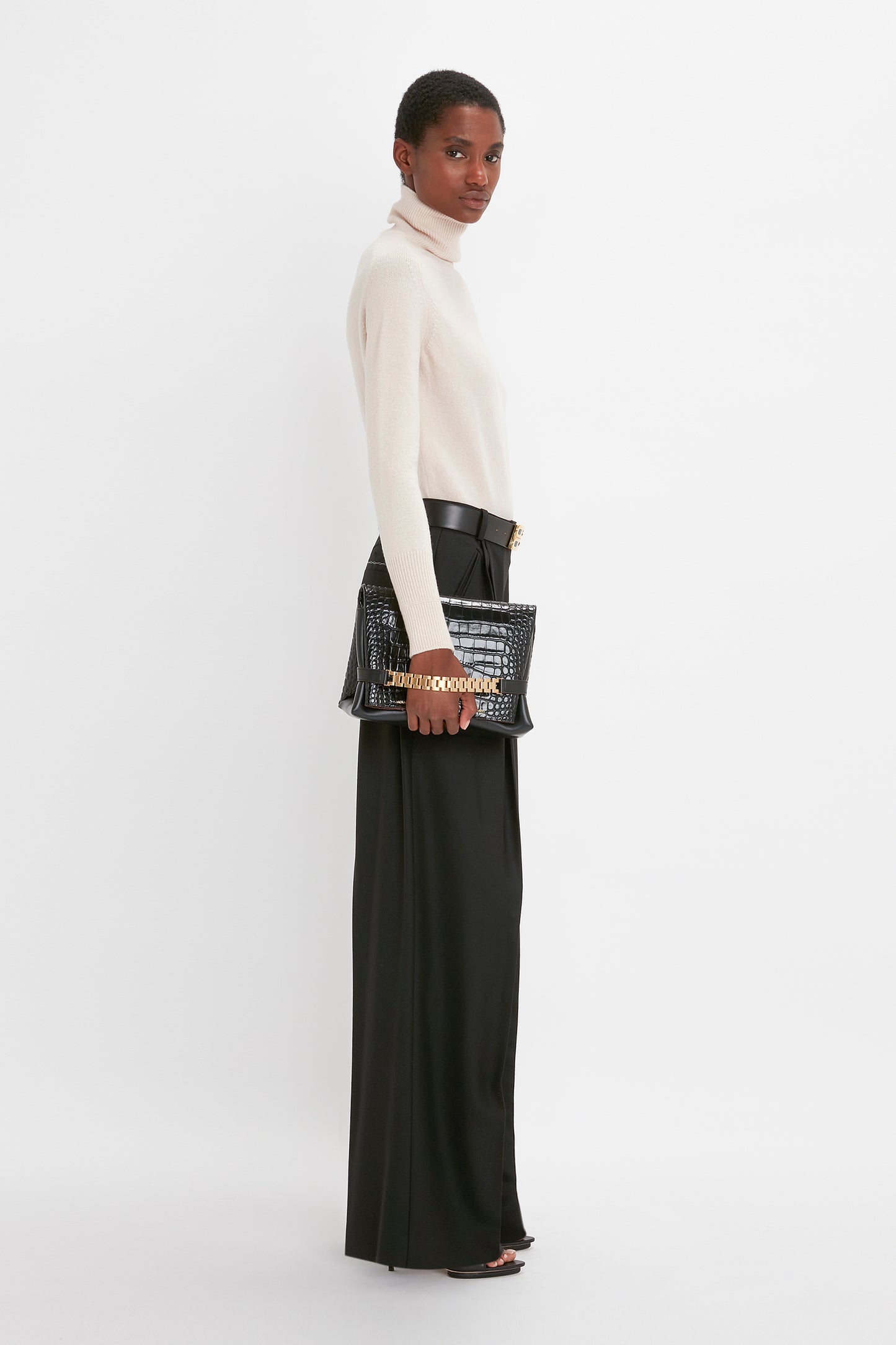 A woman in a cream lambswool Victoria Beckham polo neck sweater and black trousers holds a studded black handbag, looking over her shoulder against a white background.