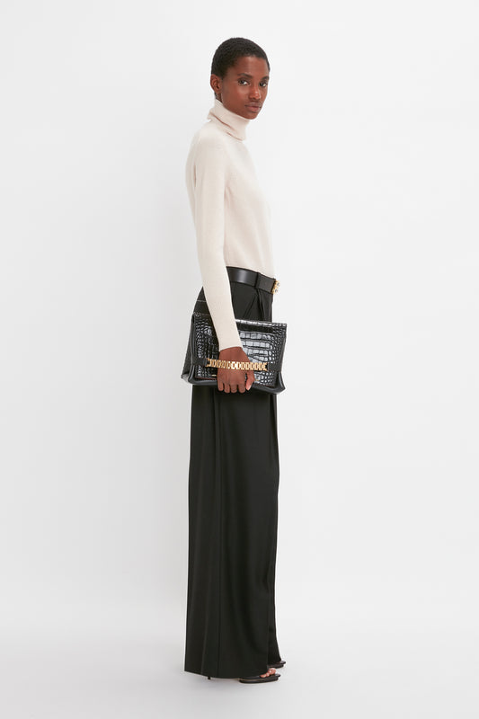 A woman in a cream lambswool Victoria Beckham polo neck sweater and black trousers holds a studded black handbag, looking over her shoulder against a white background.