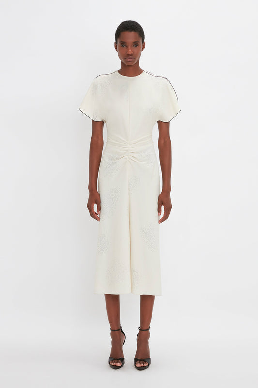 A woman stands facing forward, wearing a Victoria Beckham white embroidered gathered waist midi dress in cream with short sleeves and black strappy heels, on a plain white background.