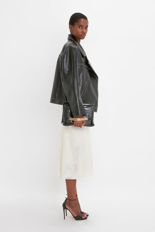 A woman in an oversized leather jacket and white lace skirt poses, looking over her shoulder in a minimalistic white studio setting.