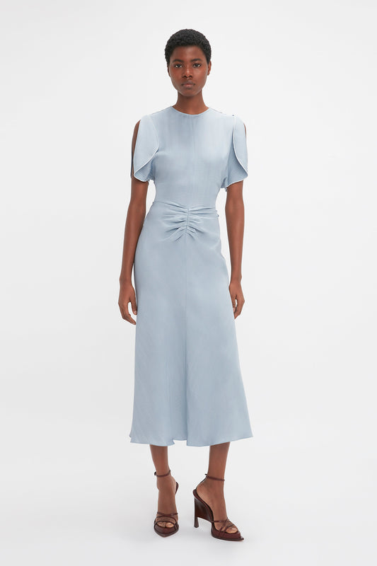 A woman stands against a white background, wearing an Exclusive Gathered Waist Midi Dress In Pebble by Victoria Beckham with cap sleeves and a front tie detail, paired with brown high heels.