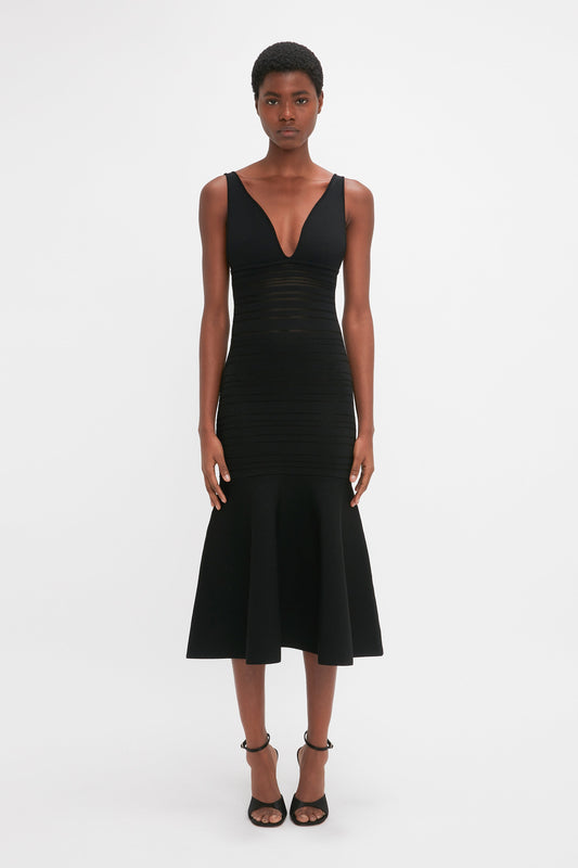 A woman in a Victoria Beckham sleek black Frame Detail Sleeveless Dress, with v-neckline and flared skirt, standing against a white background.