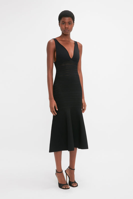 A woman in a Victoria Beckham Frame Detail Sleeveless Dress In Black with a plunging neckline and midi-length hemline, standing against a white background.