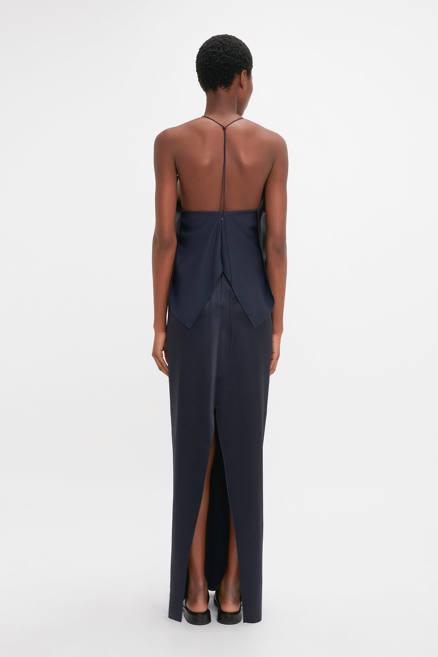 A woman wearing an elegant Victoria Beckham Frame Detail Cut-Out Cami Dress in Navy stands with her back to the camera on a white background.