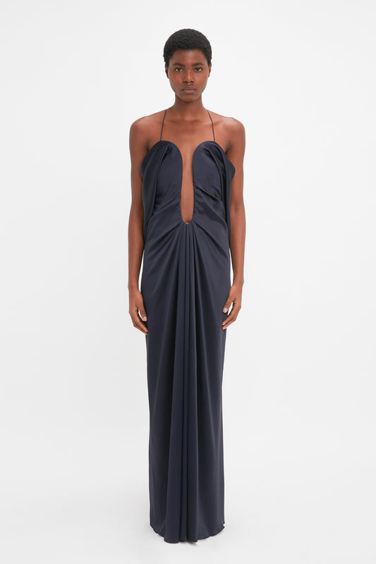 A young woman stands facing the camera, wearing an elegant evening gown in navy blue crepe back satin with a plunging neckline and draped details from the Victoria Beckham Frame Detail Cut-Out Cami Dress In Navy.