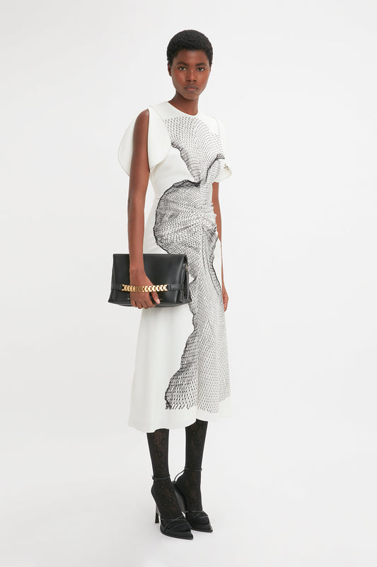 A black woman models a stylish, asymmetrical black and white Gathered Waist Midi Dress In White-Black Contorted Net by Victoria Beckham with a handbag, posing in a white studio setting.