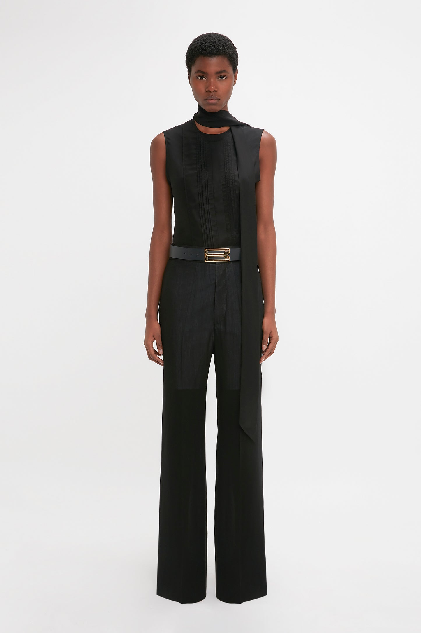 A black woman stands against a white background, modeling a sleek, sleeveless black jumpsuit with a high neckline and attached waistband detail straight leg trousers in black made from featherweight wool by Victoria Beckham.