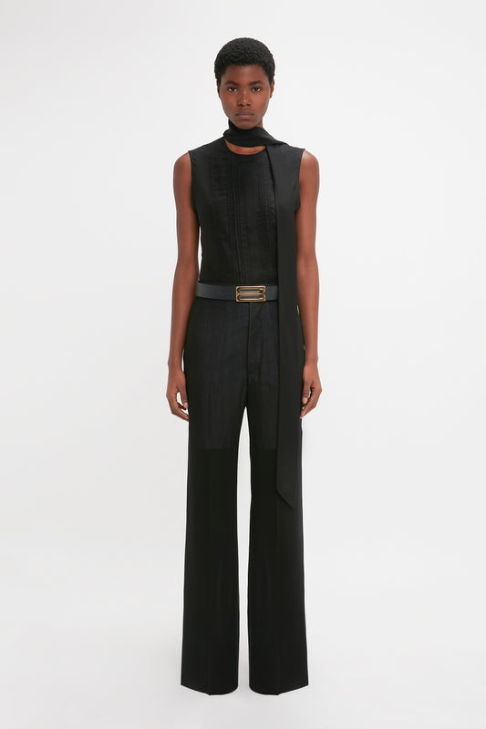 A black woman stands against a white background, modeling a sleek, sleeveless black jumpsuit with a high neckline and attached waistband detail straight leg trousers in black made from featherweight wool by Victoria Beckham.