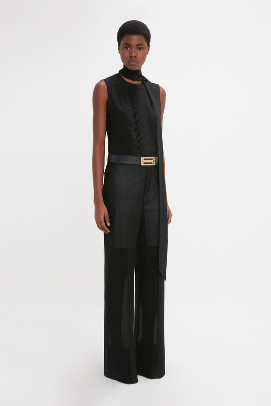 A black model standing against a white background, wearing a modern black sleeveless top, belted Victoria Beckham featherweight wool trousers with an asymmetrical overlay, and looking directly at the camera.