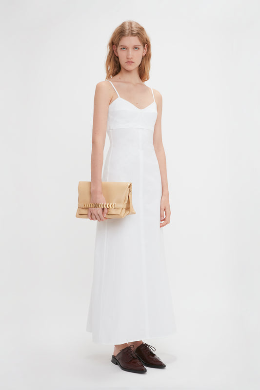 A woman in a white dress and brown shoes holds a beige Victoria Beckham Chain Pouch With Strap In Sesame Leather with a pleated design, standing against a plain white background.